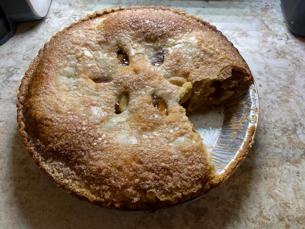A baked pie with a golden, sugared crust sits on a countertop, with one large slice missing, revealing a brown filling inside. The pie is in a disposable foil pan.