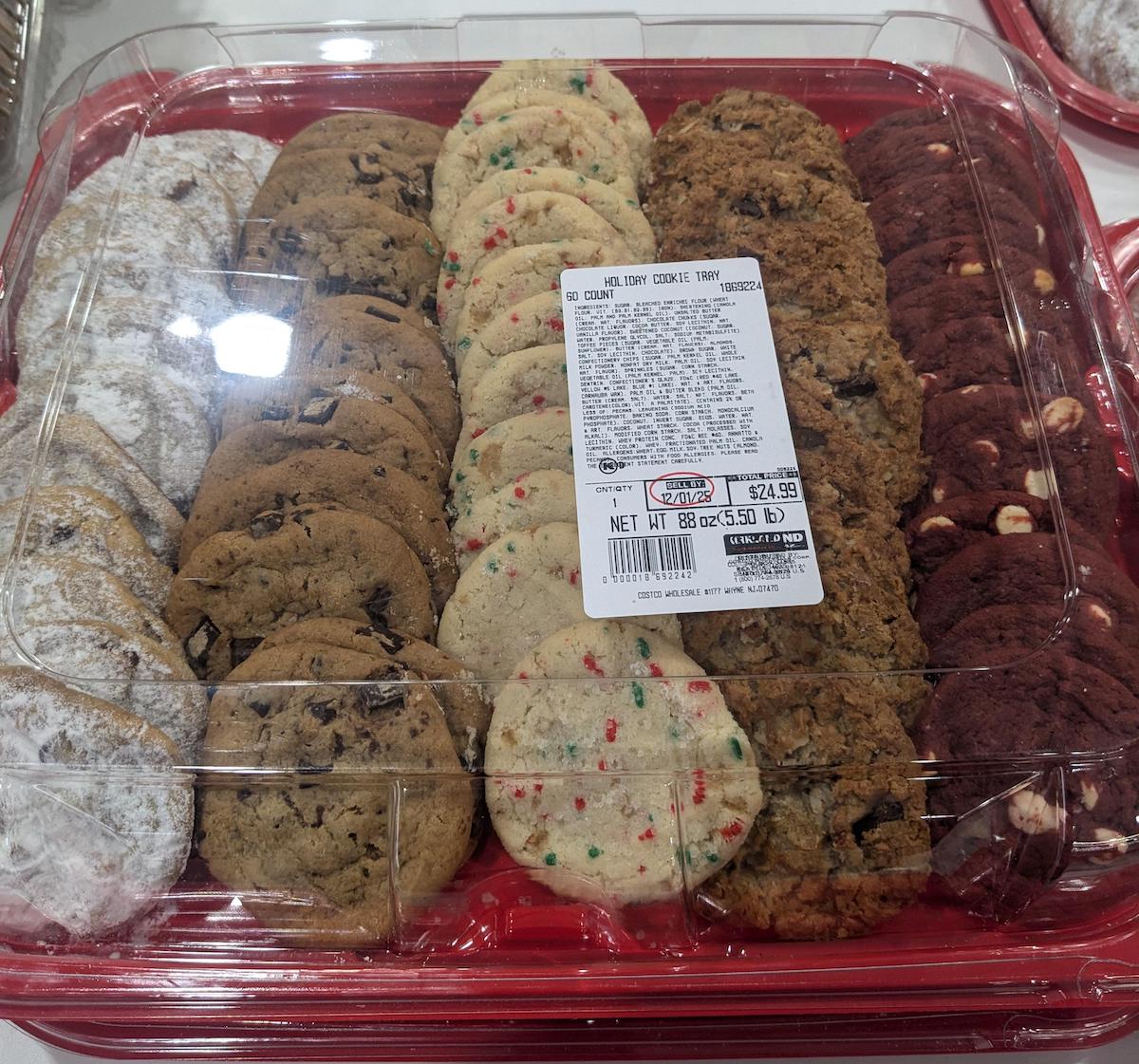 A plastic tray containing assorted cookies, including chocolate chip, powdered sugar, confetti, oatmeal, and double chocolate with white chips. A label with product and price details is placed on top of the tray.
