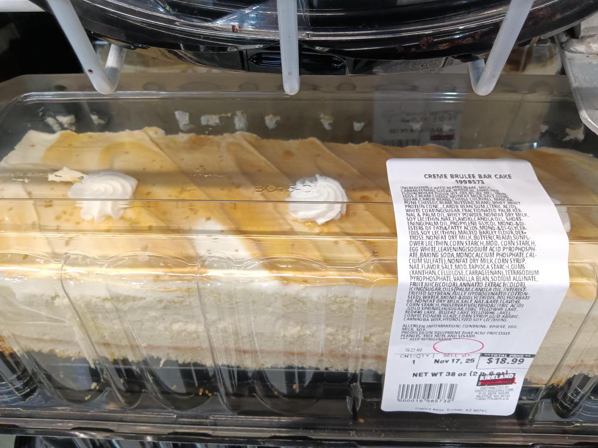 A plastic container of crème brûlée bar cake with caramel topping and two whipped cream dollops, labeled with ingredients, price, and weight, displayed on a store shelf.
