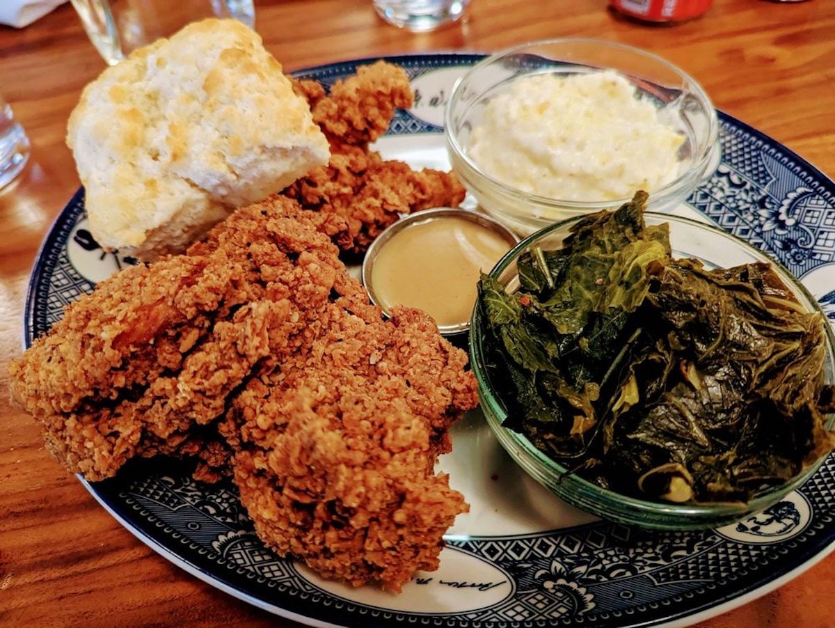 A plate with crispy fried chicken tenders, a biscuit, collard greens, mashed potatoes, and a small container of brown gravy—classic comfort food often found at charming under-the-radar restaurants—served on a patterned blue-and-white plate.