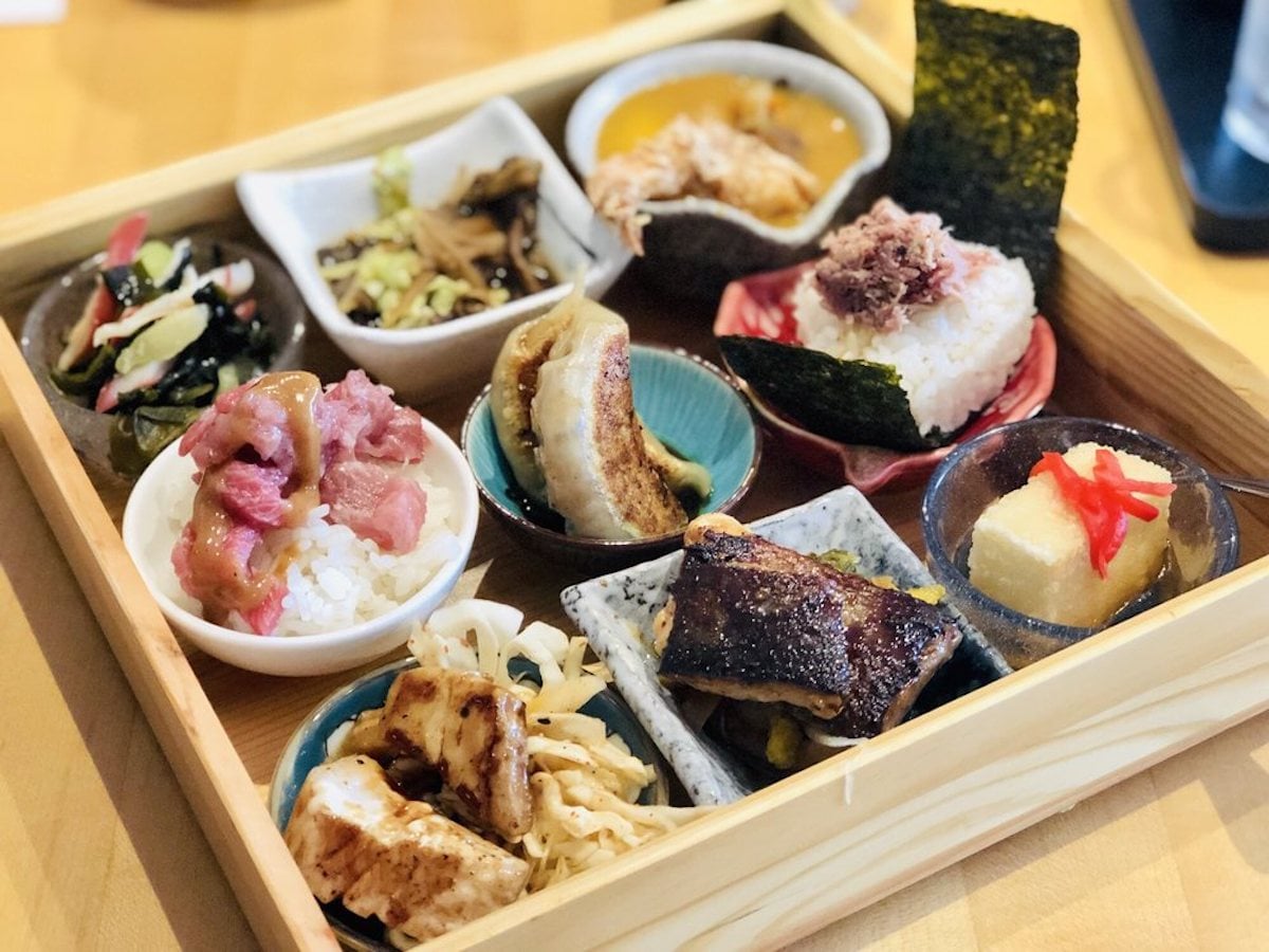 A wooden tray filled with assorted Japanese dishes in small bowls, like rice balls, grilled fish, gyoza dumplings, tofu, and sides—just like you'd find at under-the-radar restaurants known for colorful, authentic meals.
