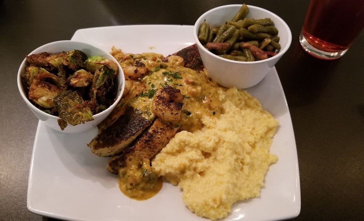 A square white plate with grilled fish topped with creamy sauce and shrimp, served with yellow grits. On the side are bowls of roasted Brussels sprouts and green beans—a feast often found at under-the-radar restaurants. A glass of red drink is in the background.