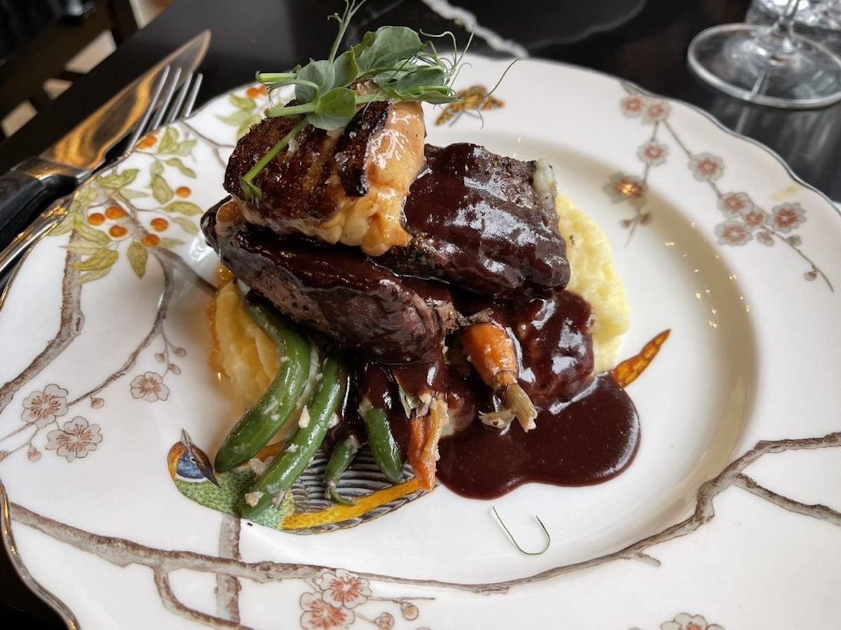 Plated gourmet dish with glazed beef topped with sauce, served over mashed potatoes and green beans on a decorative floral-patterned plate, garnished with fresh greens—a signature from one of the city’s best under-the-radar restaurants.