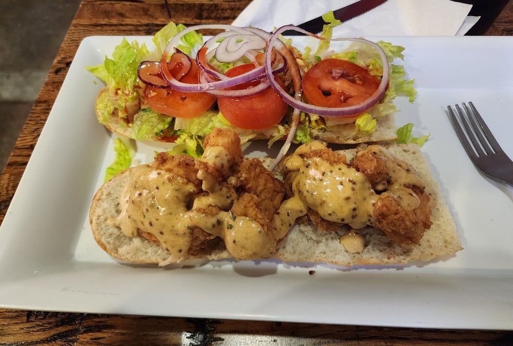 A sandwich on a white plate, topped with fried chicken, creamy sauce, shredded lettuce, tomato slices, and red onion rings—served open-faced. The kind of dish you might find at under-the-radar restaurants. A fork and napkin sit beside the plate.