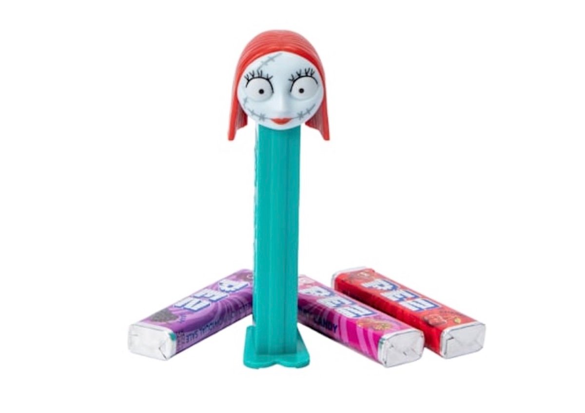 A PEZ dispenser with a red-haired character head stands upright, surrounded by three packs of PEZ candy in purple, pink, and red wrappers.