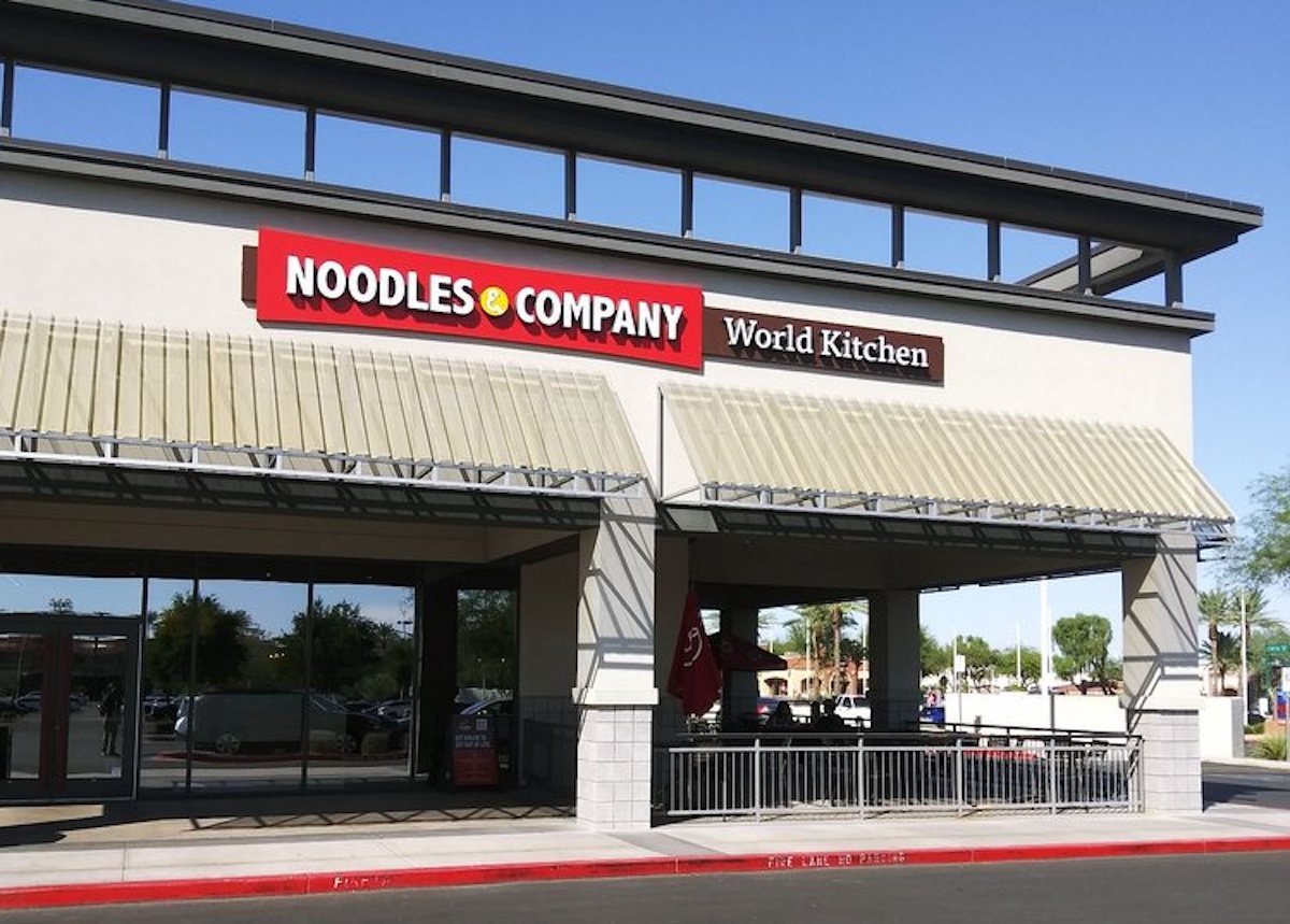 Exterior view of a Noodles & Company restaurant with a "World Kitchen" sign, outdoor seating area, awnings, large windows, and a clear blue sky in the background.
