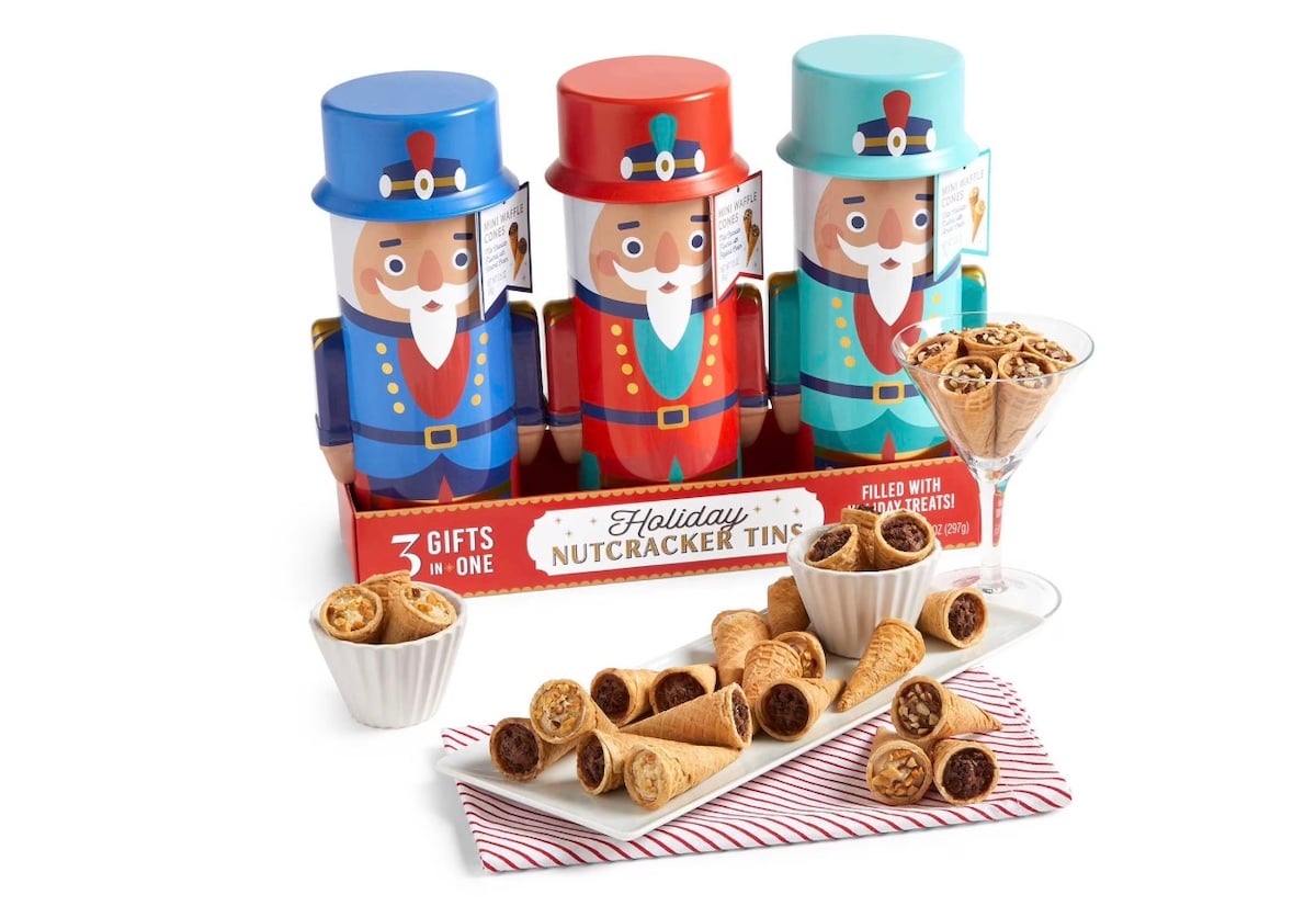 Three colorful nutcracker-themed tins filled with pastry cookies are displayed, with cookies arranged on a white plate, in small bowls, and in a martini glass in front of the tins. A red and white napkin sits under the plate.