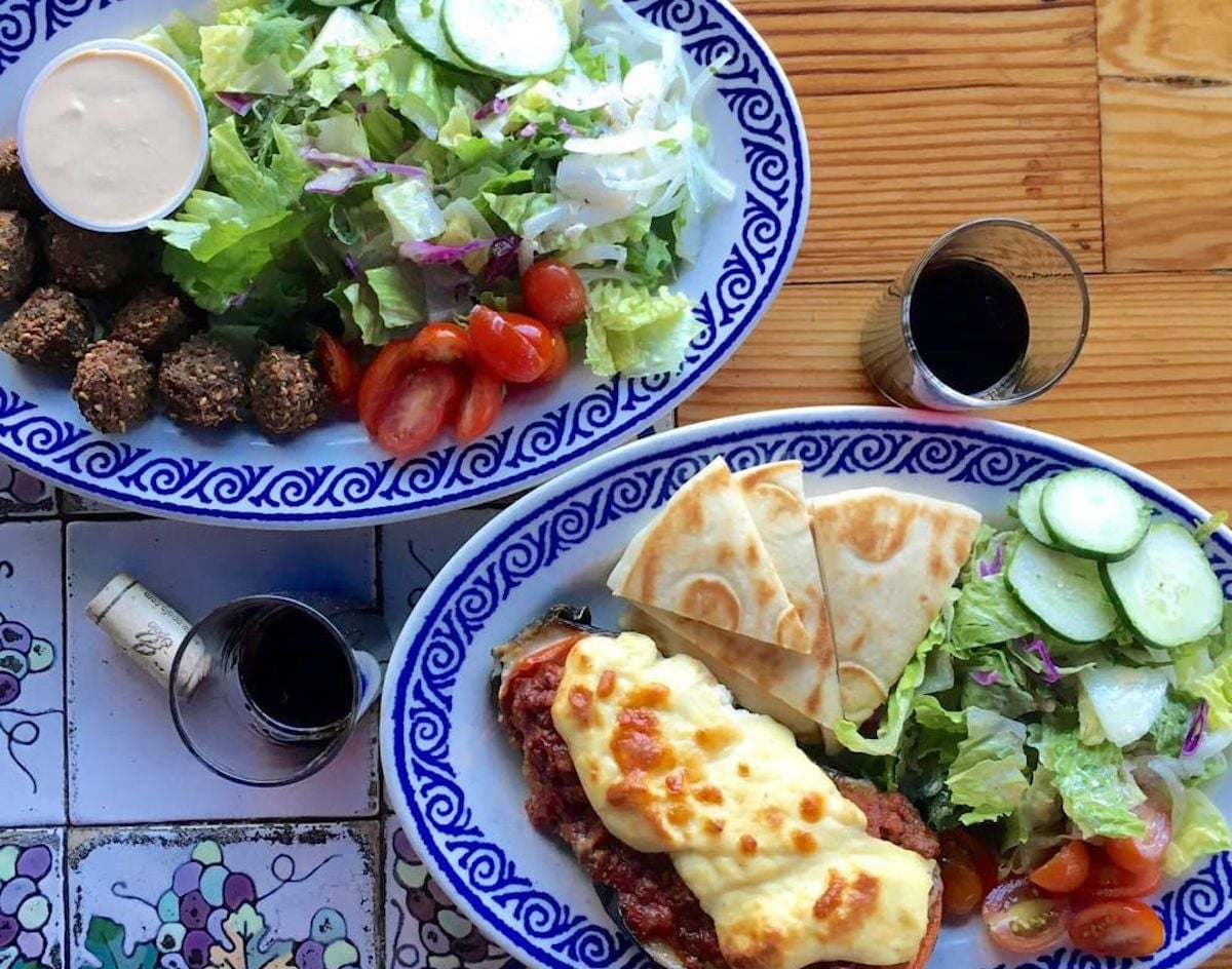 Two plates of Mediterranean food at one of the city's under-the-radar restaurants: one with salad, falafel, and dipping sauce; the other with salad, pita bread, and a cheesy baked dish. Two glasses of dark beverage and a metal cup complete the scene.