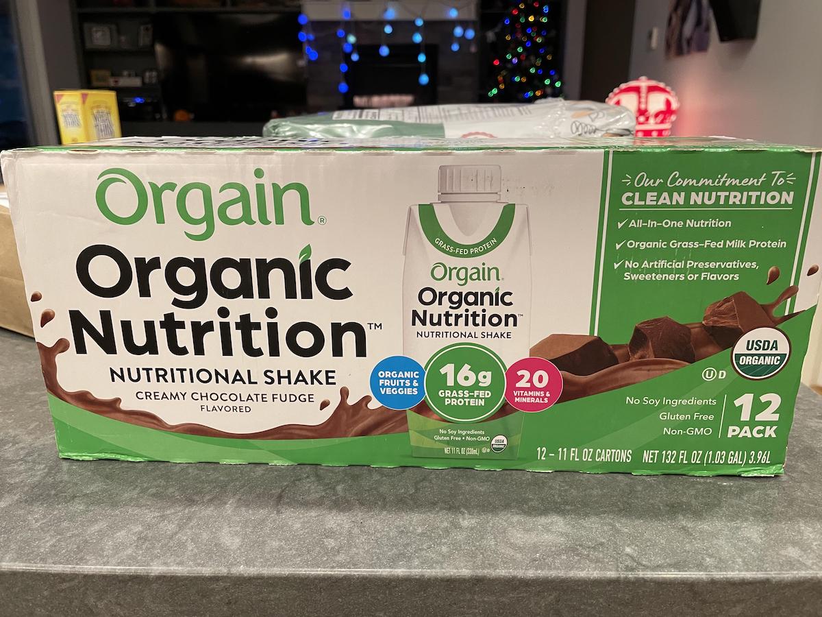 A box of Orgain Organic Nutrition Creamy Chocolate Fudge nutritional shakes sits on a counter. The packaging highlights 16g protein, 20 vitamins and minerals, and that it is organic, gluten free, and non-GMO.