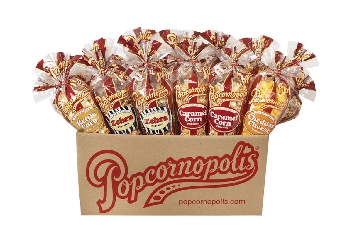 A Popcornopolis box holds six clear bags of popcorn, each labeled with a flavor: Kettle Corn, Zebra, Caramel Corn, and Cheddar Cheese. The bags are tied with red and gold twist ties.
