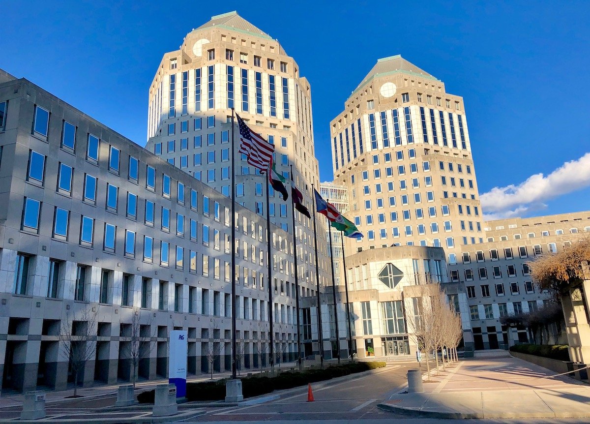 Two tall, modern office buildings with many windows stand side by side. Several flags, including the American flag, are displayed in front. The sky is clear and blue, and sunlight casts shadows on the plaza below.