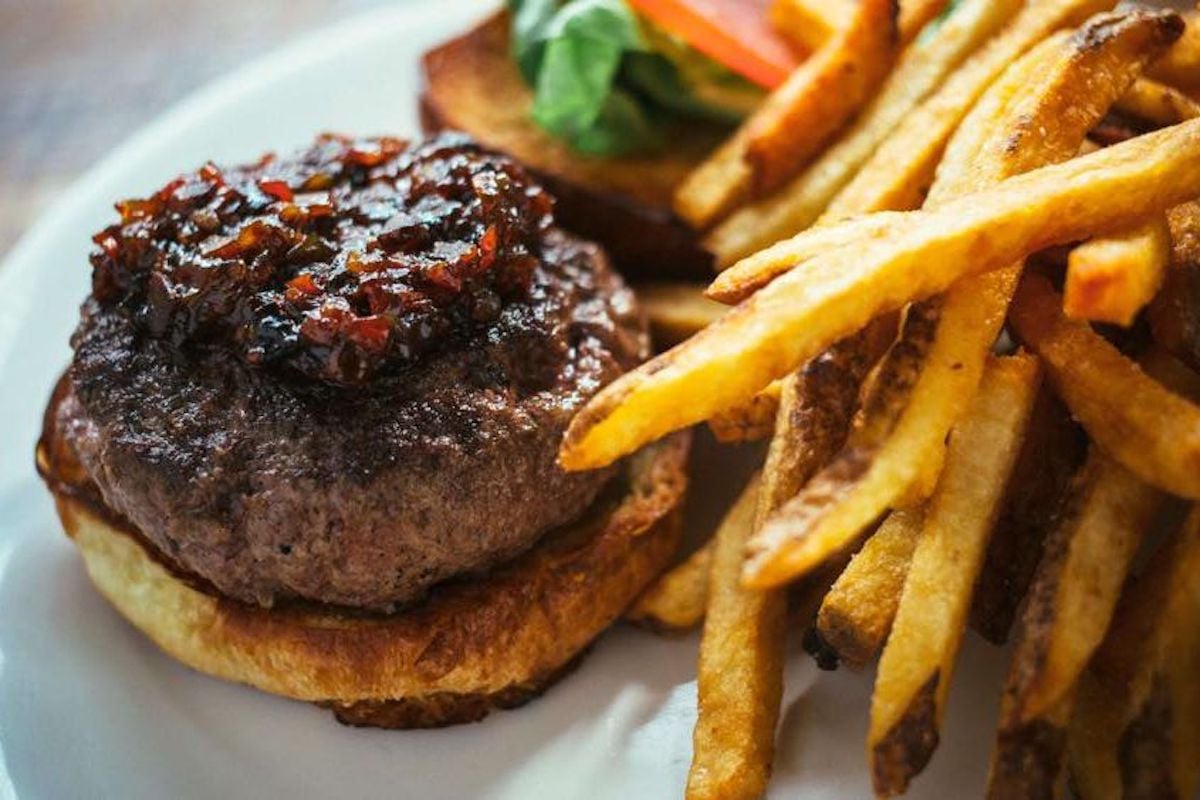 A close-up of a hamburger patty topped with relish on a toasted bun, served with golden French fries. Sliced tomato and lettuce are visible in the background—a classic find at under-the-radar restaurants.