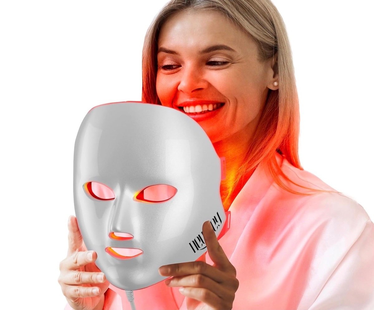 A smiling woman wearing a pink robe holds a white LED light therapy mask emitting a red glow, with the light illuminating her neck and chin.