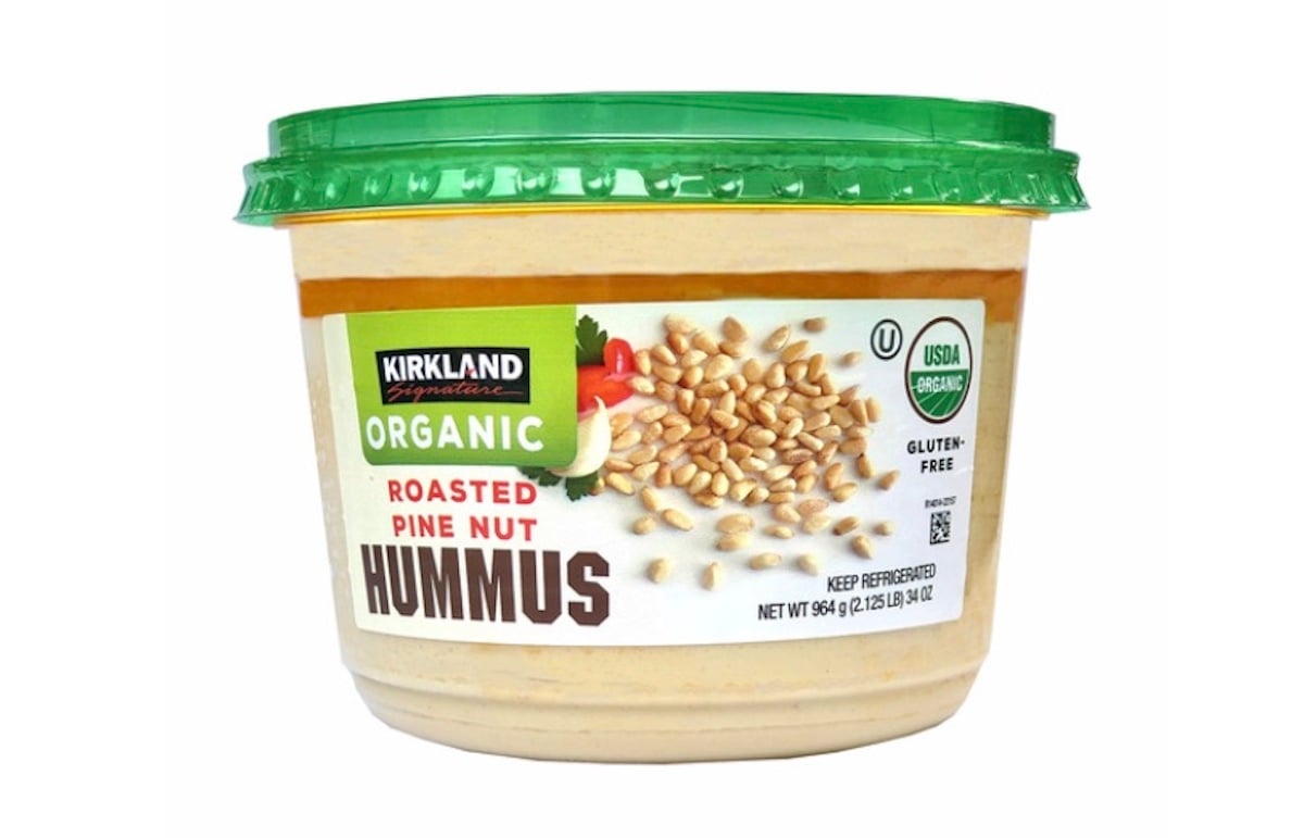 A container of Kirkland Signature Organic Roasted Pine Nut Hummus with a green lid, showing chickpeas and pine nuts on the label. The packaging displays “USDA Organic,” and “Gluten Free.”