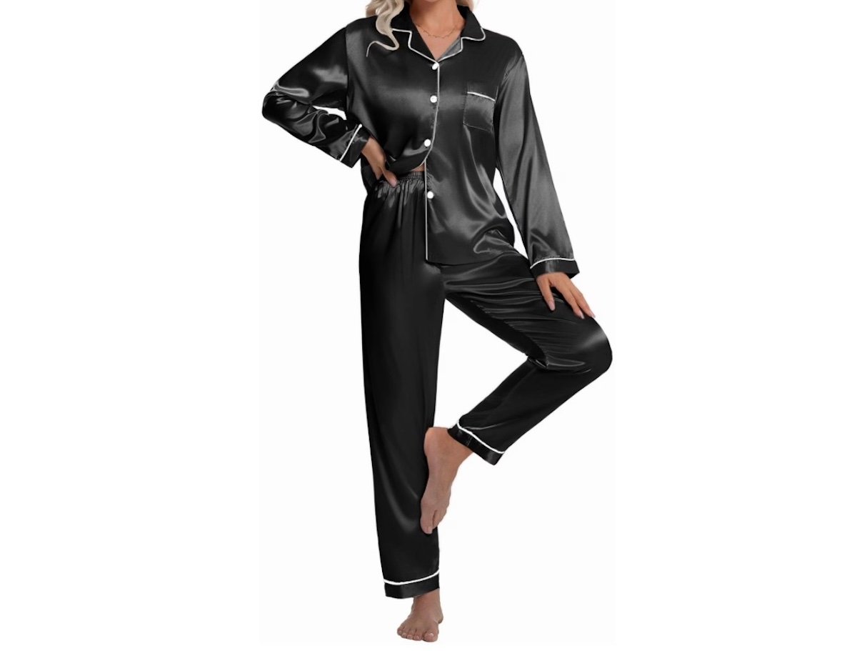 A woman wearing a black satin pajama set with white piping, featuring a long-sleeve button-up top and matching pants, standing barefoot on one leg against a white background.