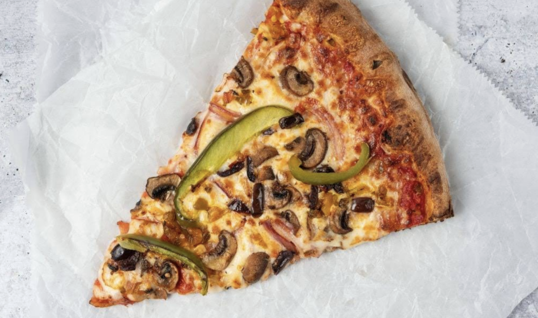 A slice of east coast cheap pizza with mushrooms, green bell peppers, and melted cheese on parchment paper, set on a light-colored surface.