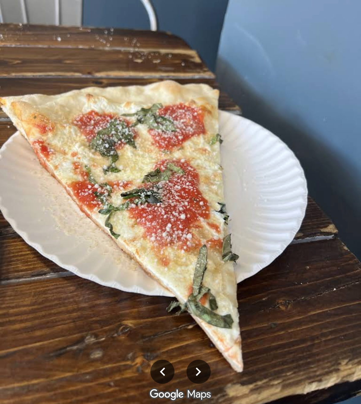 A large slice of Margherita pizza, reminiscent of classic East Coast cheap pizza slices, with tomato sauce, mozzarella cheese, fresh basil, and grated parmesan sits on a white paper plate atop a rustic wooden table.