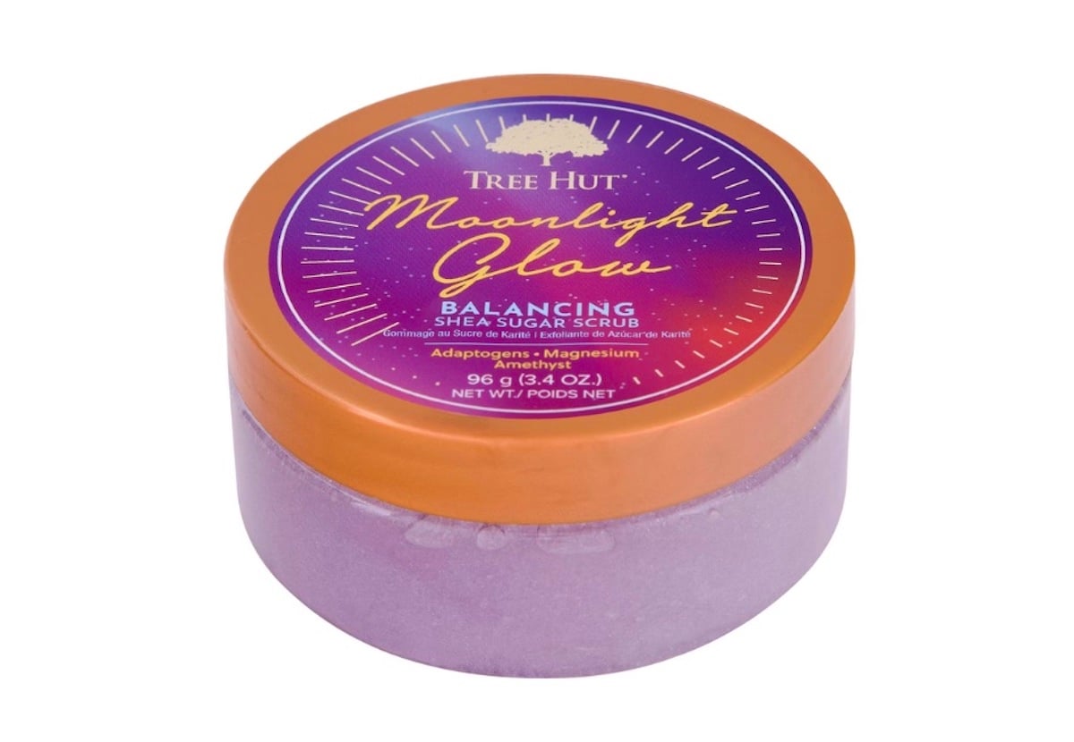 A round container of Tree Hut Moonlight Glow Balancing Shea Sugar Scrub with a purple base and an orange lid featuring product details in gold, white, and pink text. The scrub inside is a light purple color.