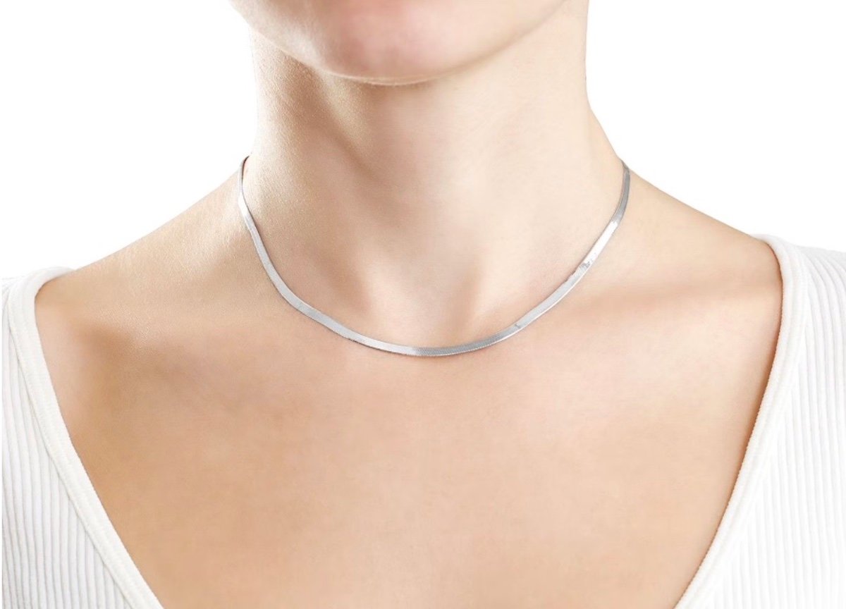 A woman wearing a thin, flat silver choker necklace and a white ribbed top, shown from the shoulders up against a plain white background.