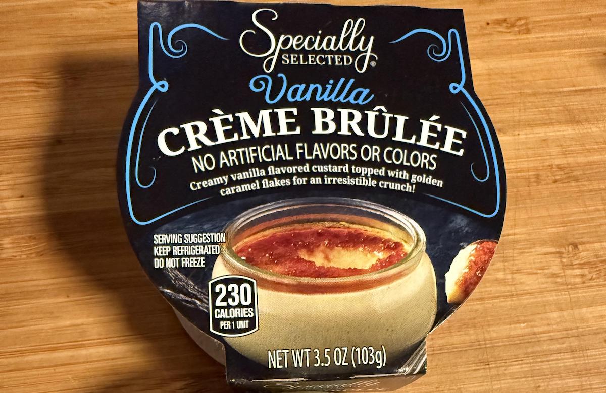 A container of Specially Selected Vanilla Crème Brûlée sits on a wooden surface. The packaging highlights no artificial flavors or colors and shows an image of the custard dessert topped with caramelized sugar.