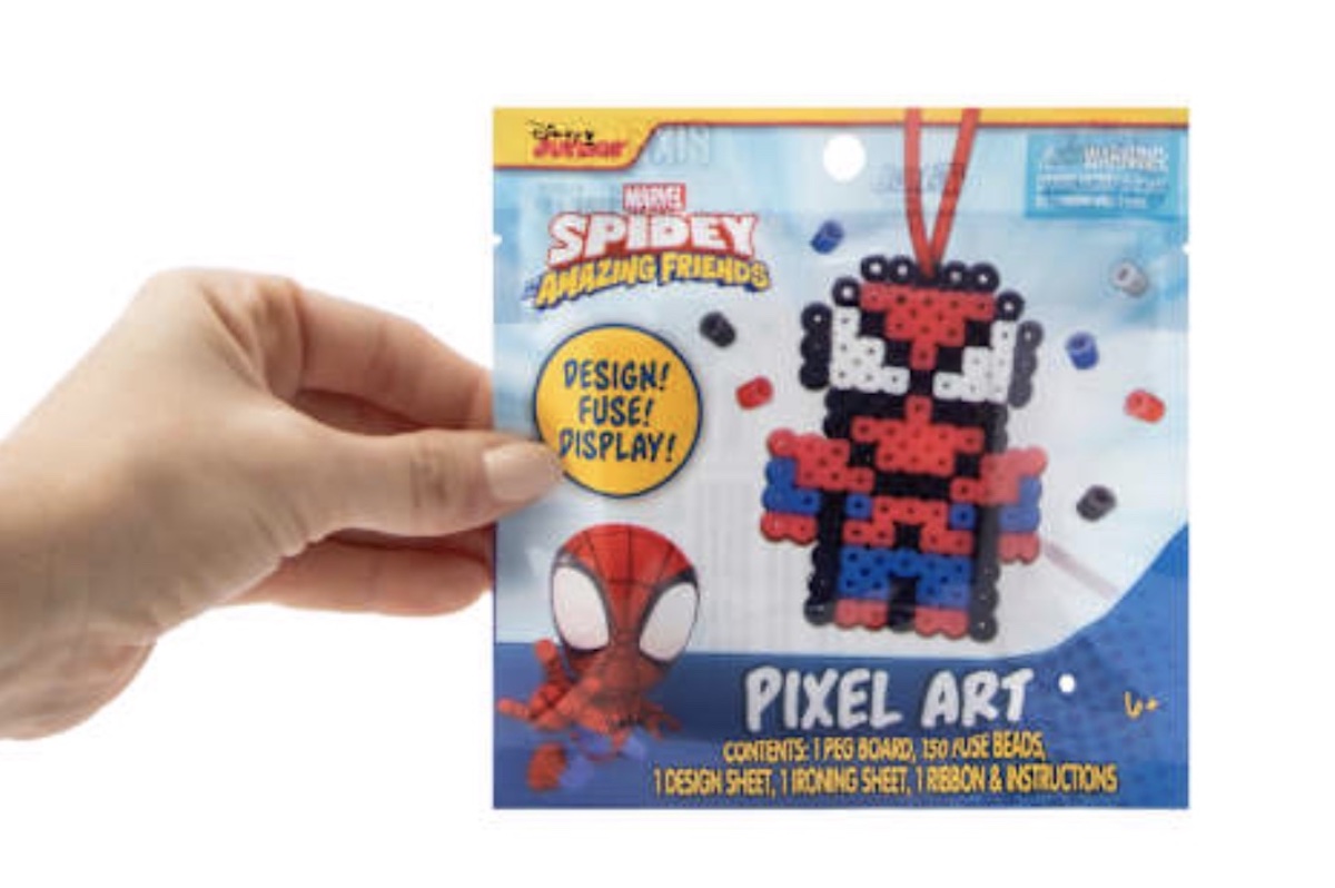 A hand holds a Spidey and His Amazing Friends Pixel Art kit, featuring a bead design of Spidey. The packaging is blue with a cartoon Spidey, and includes beads, a design sheet, ribbon, and accessories.
