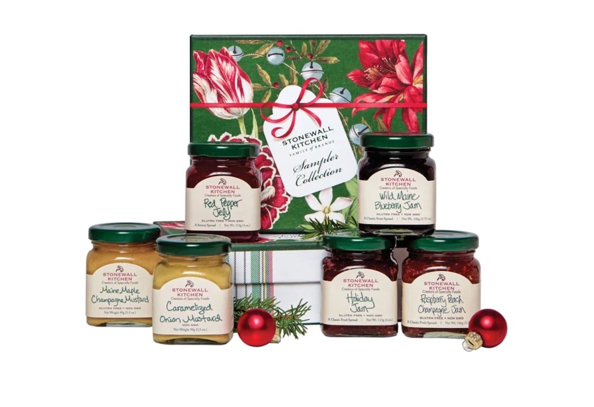 A festive gift set with a floral box and six Stonewall Kitchen jars, including jams, jellies, and mustards, arranged with small holiday ornaments and greenery.