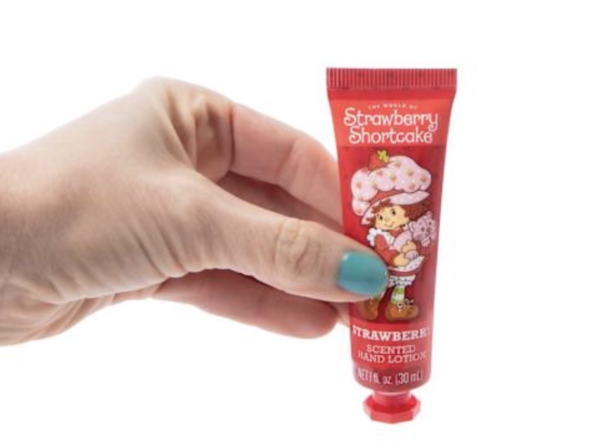 A hand with turquoise nail polish holds a small red tube of Strawberry Shortcake scented hand lotion featuring a cartoon girl with a strawberry hat on the packaging.