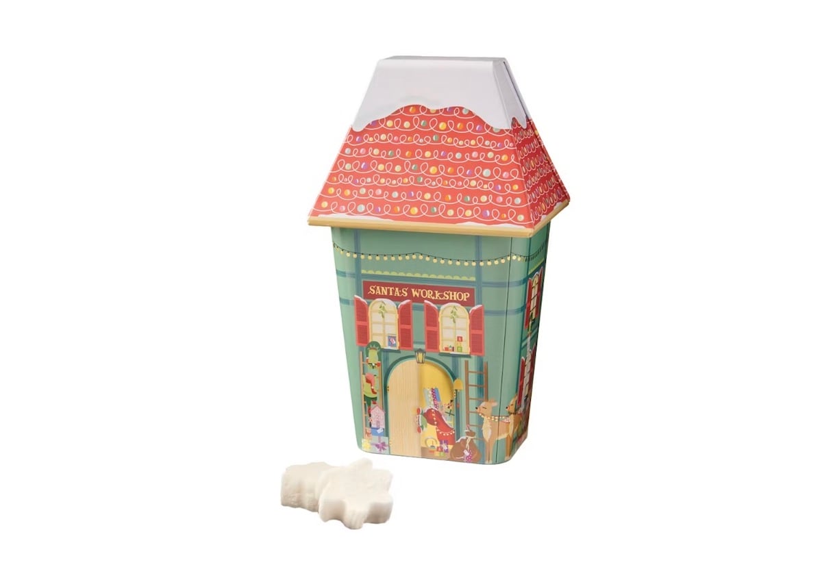 A festive, house-shaped tin decorated as Santa's Workshop with a snowy roof, colorful walls, and holiday illustrations. In front of the tin are two white bear-shaped candies or soaps.