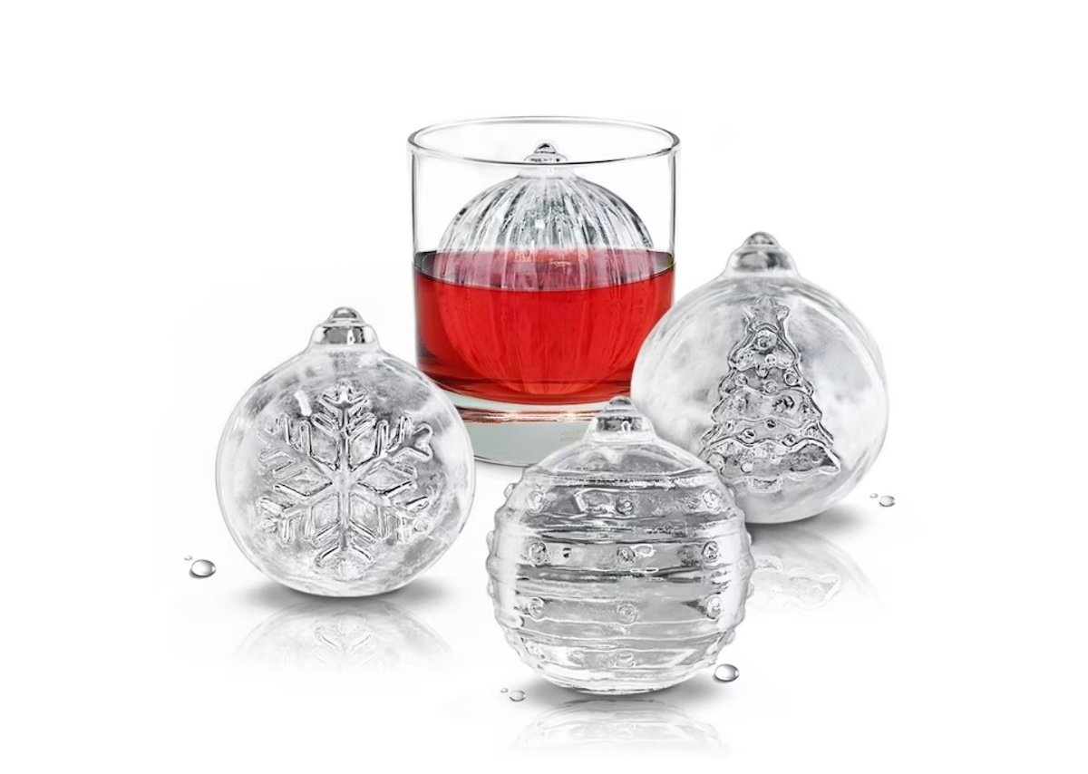 Three large, round ice spheres with holiday designs—a snowflake, stripes, and a Christmas tree—sit next to a glass of red liquid containing a similar ice sphere, all on a white background.