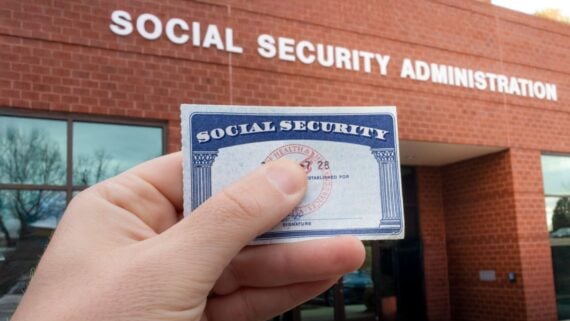 A hand holding a Social Security card in front of the Social Security Administration building with large white letters on a red brick facade. iStock-2203057418