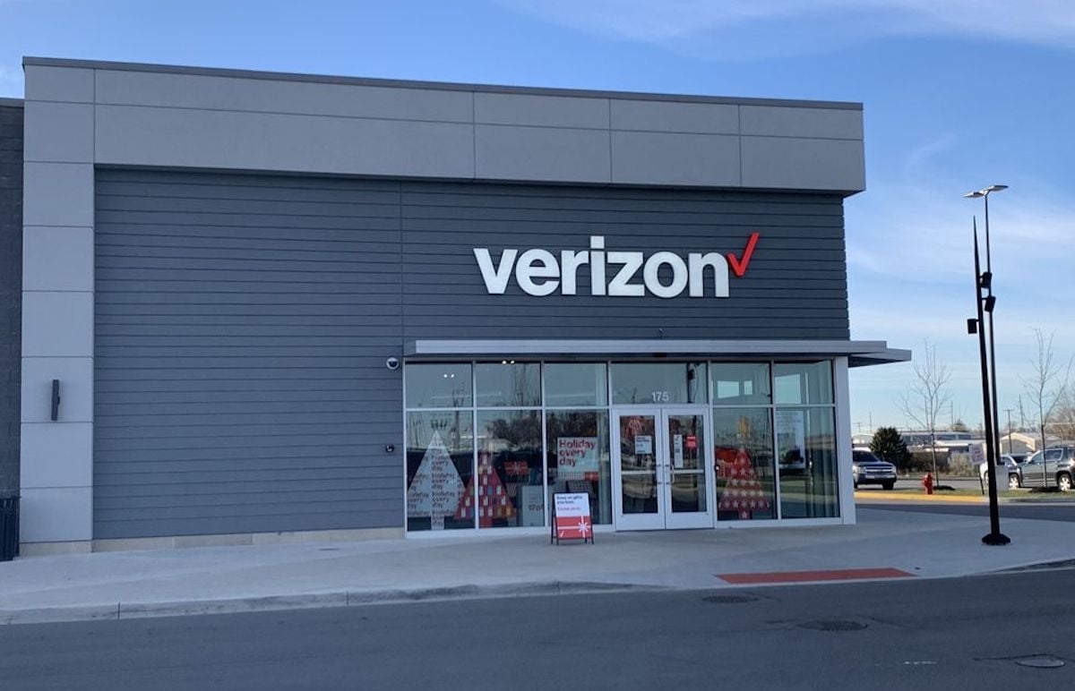A Verizon store with a gray exterior, large windows, and the Verizon logo above the entrance. The sky is clear, and there are cars and trees in the background.