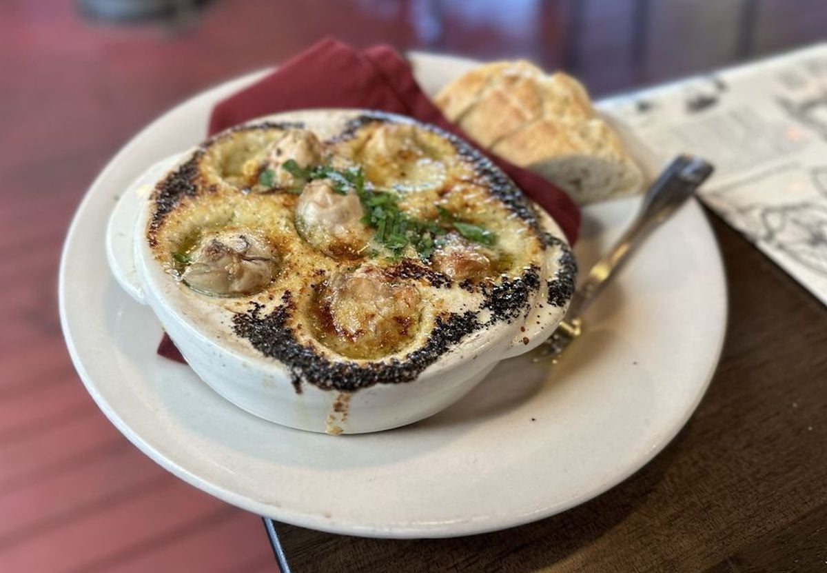 A white dish of baked escargot topped with herbs and cheese sits on a plate with a red napkin, accompanied by slices of bread and a fork—a hidden gem often found in under-the-radar restaurants.