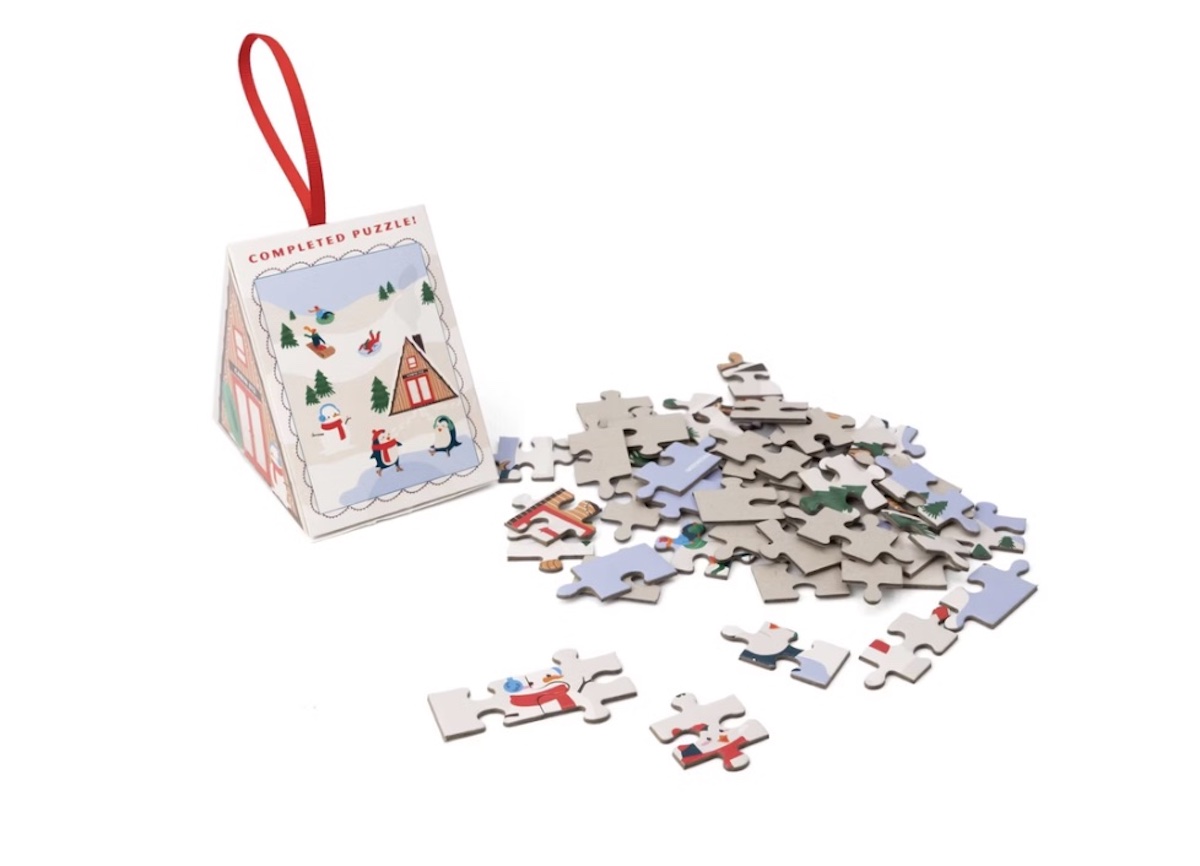 A small pile of jigsaw puzzle pieces is next to a decorative, triangular box with a red ribbon. The box shows an illustration of a winter scene with snow, trees, and people sledding and playing.