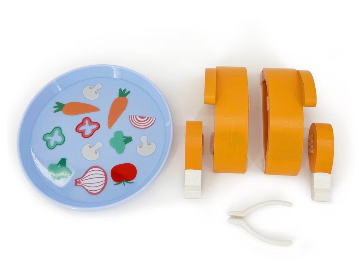 A blue plate with wooden cutouts of vegetables, including carrots, mushrooms, peppers, onion, and tomato, next to segmented orange toy pieces and a small white tong on a white background.