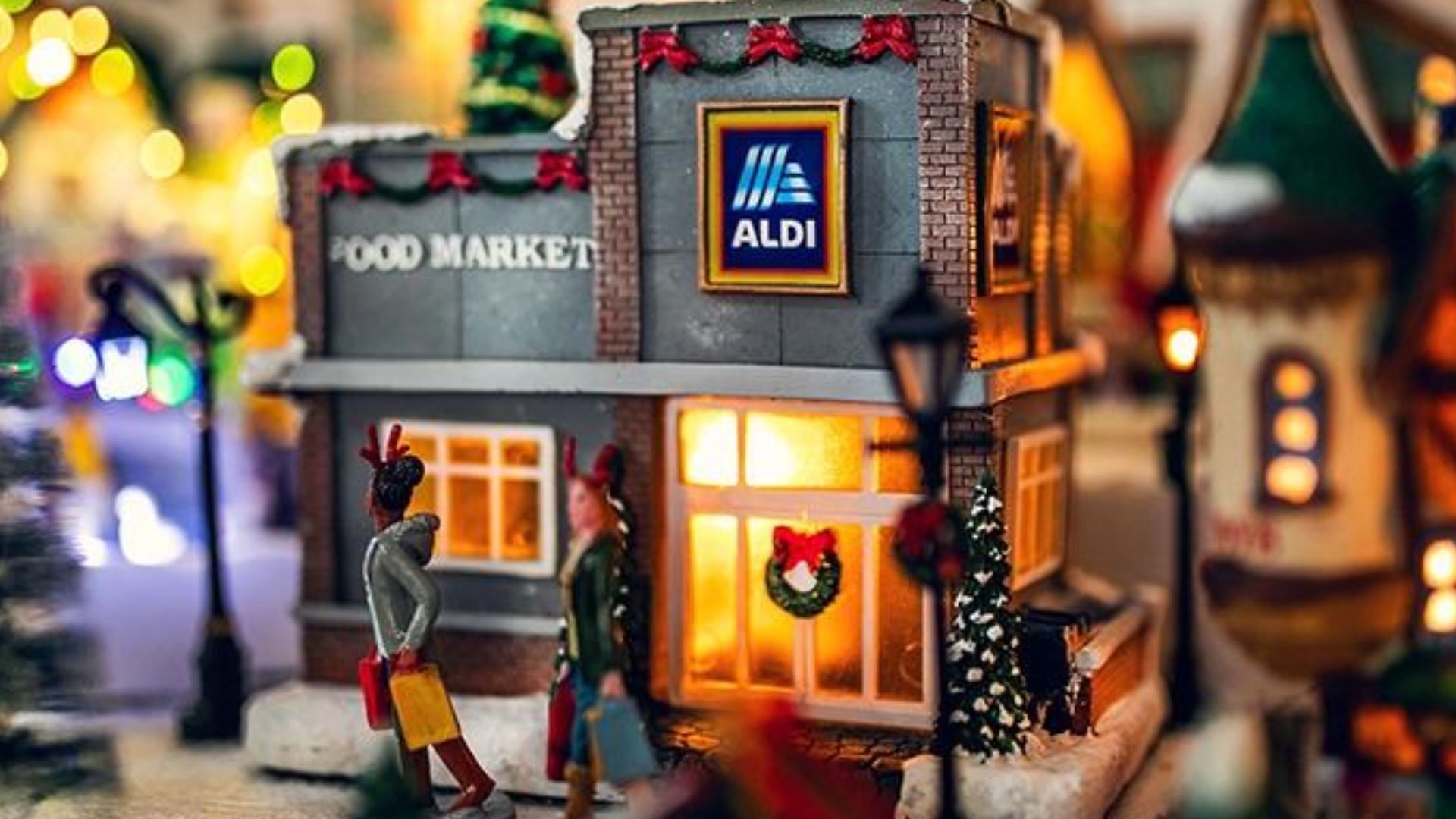 A festive miniature model of an ALDI food market decorated with wreaths and garlands. Warm lights glow inside as two small figurines with shopping bags walk past the building.