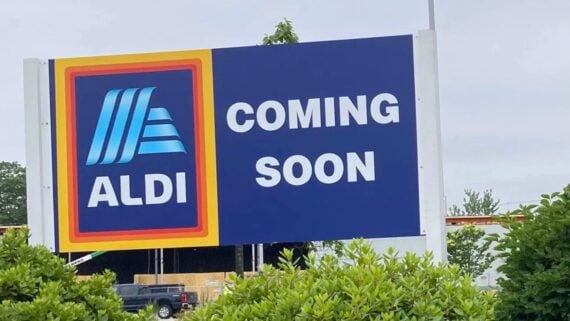 A large ALDI sign with the text "COMING SOON" stands in front of bushes, indicating the store's future opening. A partial view of a building and parked truck are visible in the background.