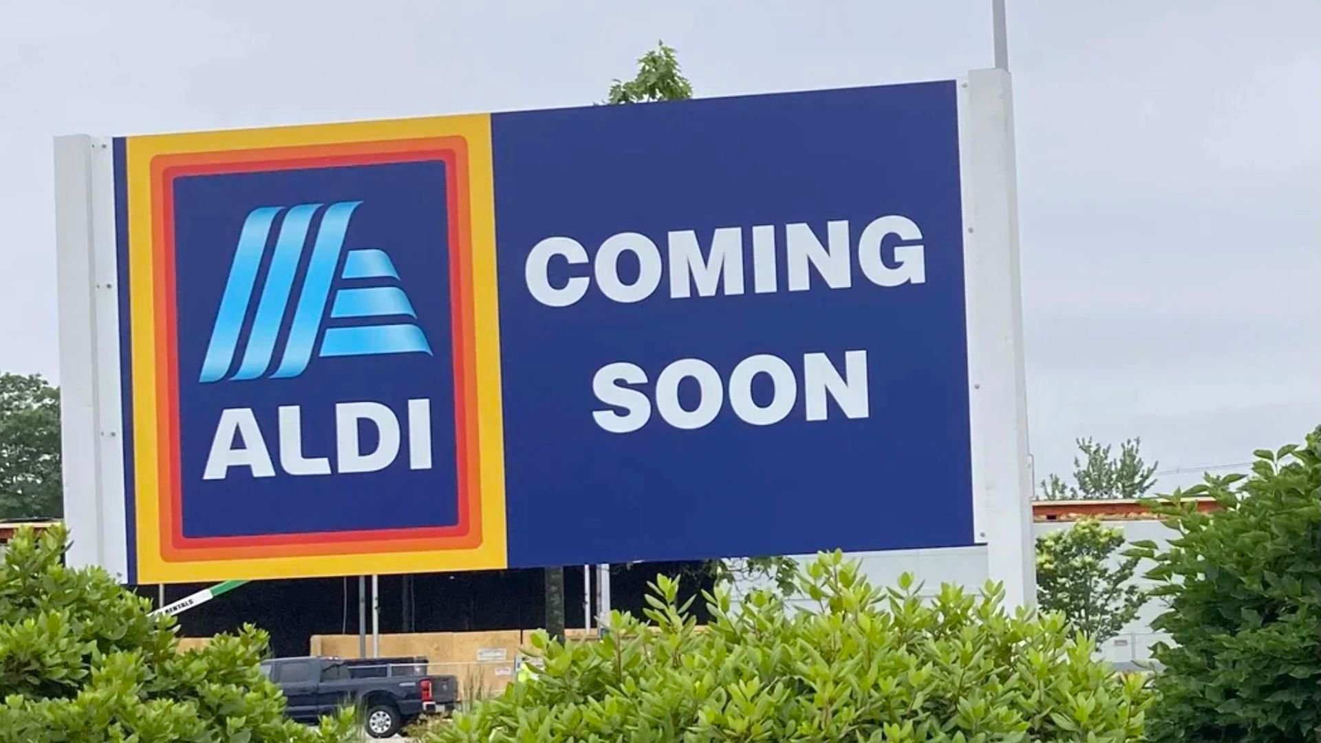 A large ALDI sign with the text "COMING SOON" stands in front of bushes, indicating the store's future opening. A partial view of a building and parked truck are visible in the background.