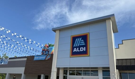 The front entrance of an ALDI grocery store with a large ALDI logo sign, blue and white pennant flags, and a "Now open!" banner visible on the building.