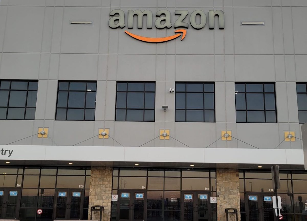 The front of an Amazon warehouse building with large Amazon logo above a row of windows and glass entrance doors below.