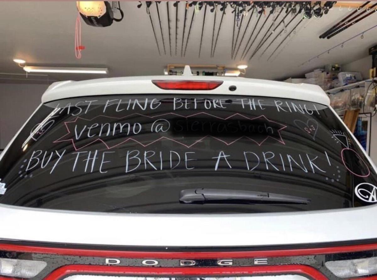 A white Dodge SUV’s rear window decorated with handwritten messages reads: “Last fling before the ring! Venmo @sierrasbach. Buy the bride a drink!” The car is parked in a garage with fishing rods on the ceiling.