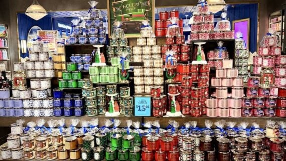 A store display features large, colorful stacks of candles in decorated jars arranged by color, with gift sets on the bottom shelf and a holiday sign above. The sign advertises a candle sale for $13.95 each.