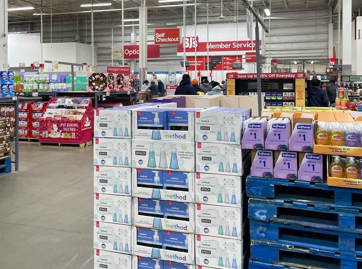 A warehouse store interior with stacked boxes of cleaning products in the foreground, displays of goods on shelves, and shoppers near checkout and service counters in the background. Signs for self-checkout and member service are visible.