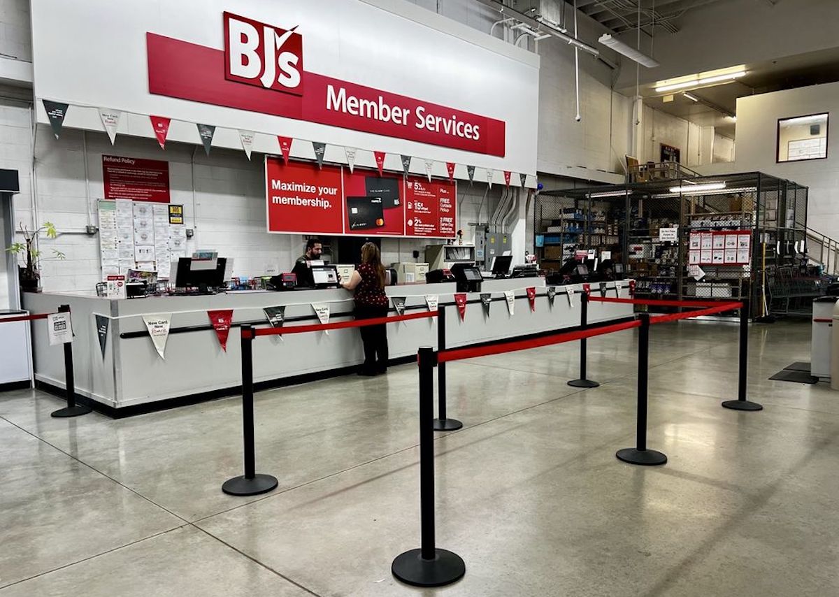 BJ’s Member Services counter inside a store, with one staff member behind the counter and signboards above. Red and black stanchions form a line, and shelves with products are visible in the background.