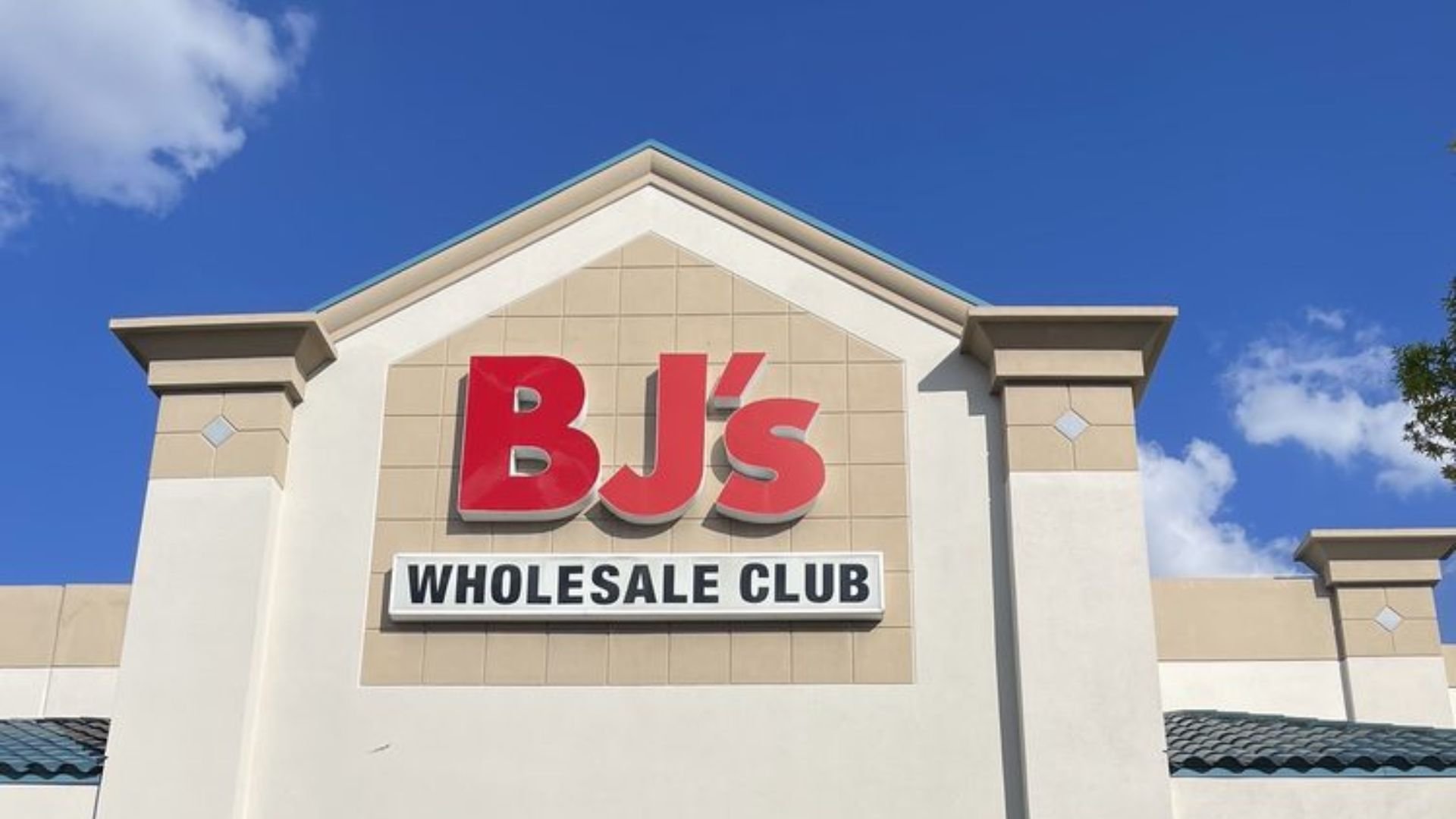 The exterior sign of BJ's Wholesale Club on a beige building, with a bright blue sky and some clouds in the background.