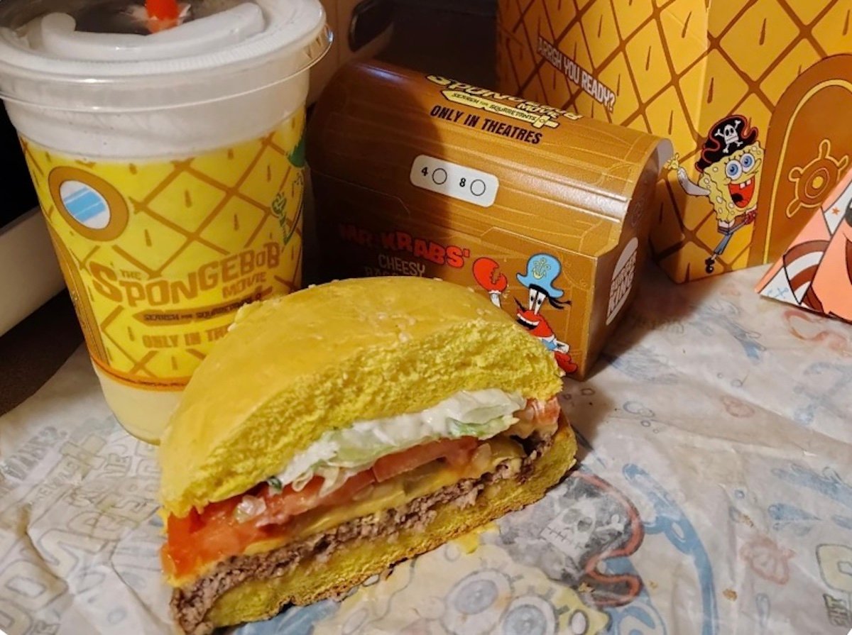 A Spongebob-themed fast food meal featuring a yellow pineapple cup, a burger with lettuce and tomato, and packaging decorated with Spongebob and Mr. Krabs graphics on a wrapper.