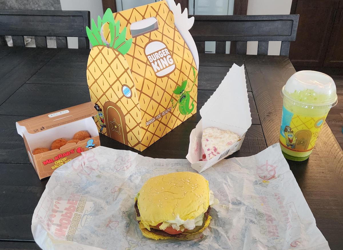 A Burger King meal themed after SpongeBob, featuring a yellow pineapple-shaped box, burger with yellow bun, nuggets, a green drink, and a cake slice on a table. Cartoon graphics decorate the packaging.
