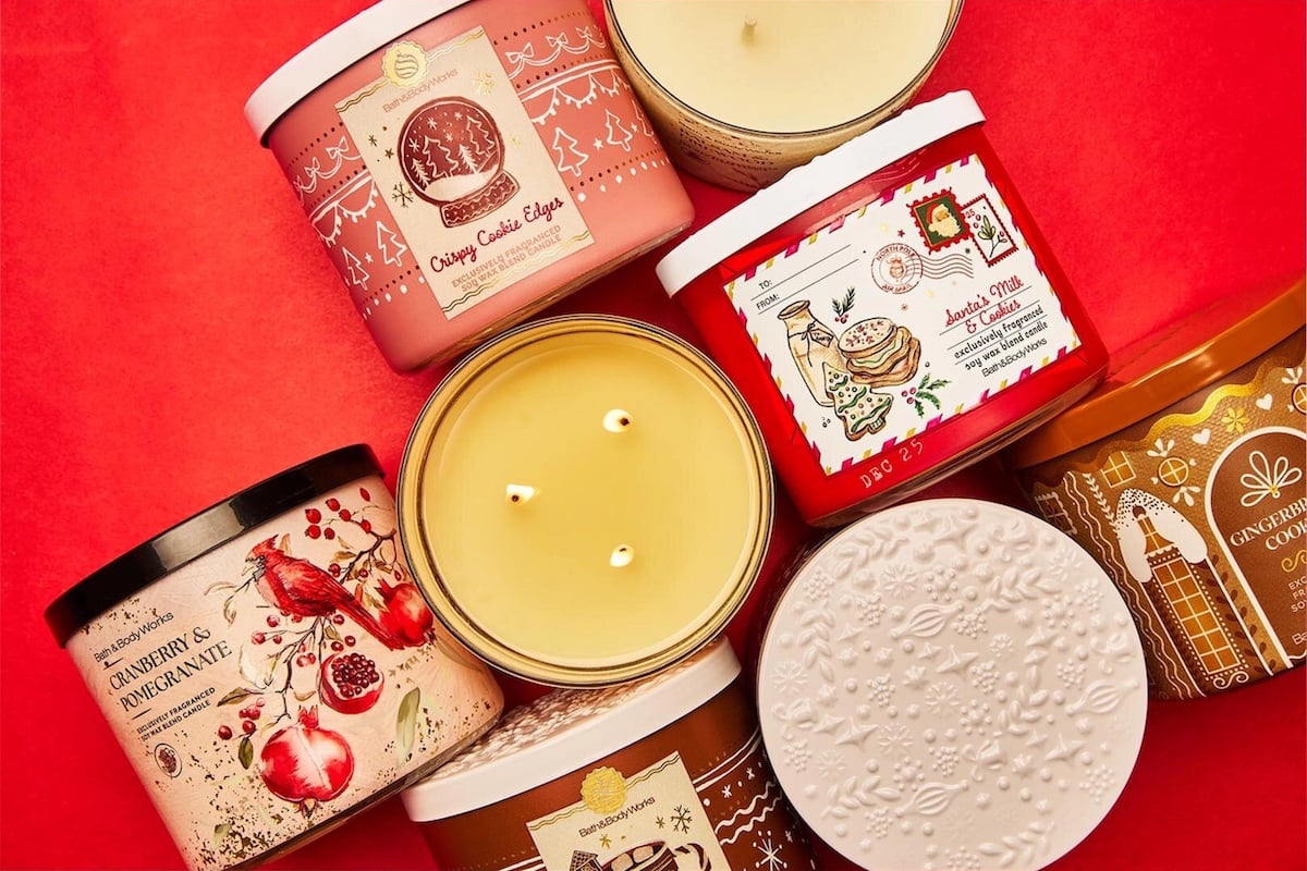 Assorted holiday-themed candles in festive jars arranged on a red background, featuring designs with gingerbread, pomegranate, cookies, and decorative patterns.