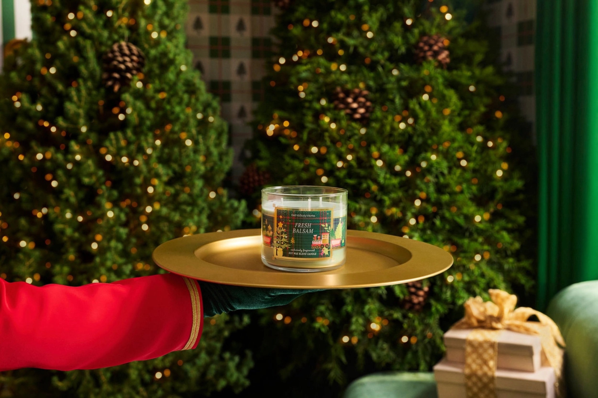 A hand in a green glove and red sleeve holds a gold tray with a holiday-scented candle in front of decorated Christmas trees and wrapped gifts.