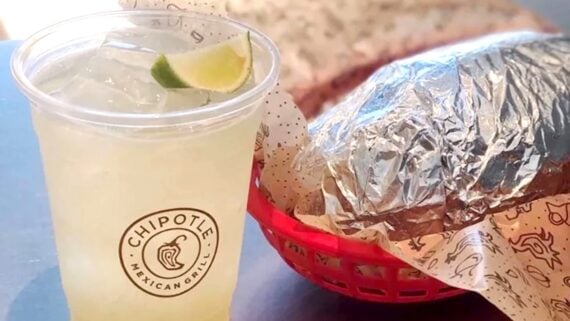 A plastic cup of lemonade with ice and a lime wedge sits next to a foil-wrapped burrito in a red basket lined with branded paper from Chipotle Mexican Grill.