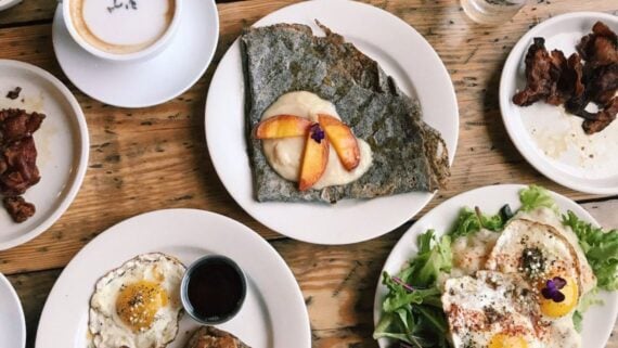 A rustic wooden table set with plates of fried eggs, bacon, a buckwheat crepe topped with fruit and cream, salad greens, toast with syrup, and a cup of coffee with foam art&mdash;just like you'd find at beloved under-the-radar restaurants.